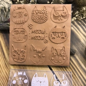 May include: A set of nine brown rubber stamps featuring different cat designs. The stamps are arranged in a 3x3 grid on a brown background. The stamps feature various cat faces, including a cat with a crown, a cat with a heart, and a cat with a paw print. The text "MEOW MEOW" is also included on the stamps.