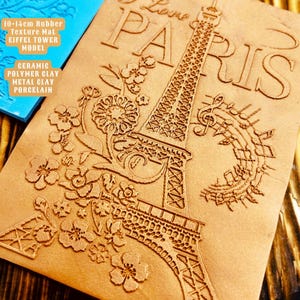 以下が含まれることがあります： エッフェル塔、花、音符のエンボス加工が施された茶色のゴム製テクスチャマット。マットには「J'aime Paris」の文字もエンボス加工されています。