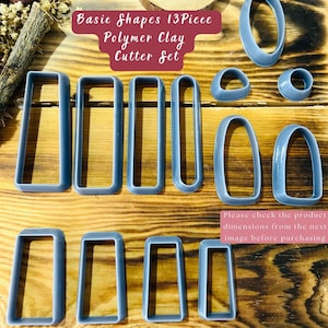 Puede incluir: Un juego de 13 cortadores de arcilla polimérica grises en varias formas rectangulares y ovaladas. Los cortadores están dispuestos sobre una superficie de madera. El texto "Basic Shapes 13 Piece Polymer Clay Cutter Set" se muestra sobre los cortadores. El texto "Please check the product dimensions from the next image before purchasing" se muestra debajo de los cortadores.