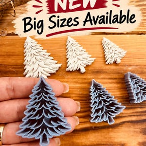 Puede incluir: Una colección de cortadores de galletas de plástico gris y blanco con forma de árbol de Navidad. Los cortadores presentan diseños de ramas detallados y superpuestos. Un cortador gris se sostiene en primer plano. El texto dice NEW Big Sizes Available.
