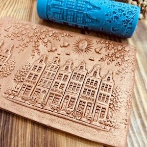 Puede incluir: Un sello rectangular de arcilla de color terracota con un diseño en relieve detallado de una hilera de casas, árboles y bicicletas. Un rodillo azul con un diseño similar de casas y árboles está encima. Para manualidades o repostería.