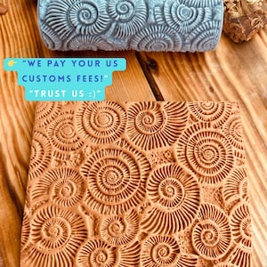 May include: A close-up of a gray roller and a brown rectangular stamp, both featuring a spiral seashell pattern. The stamp has the text "WE PAY YOUR US CUSTOMS FEES!" and "TRUST US :)".