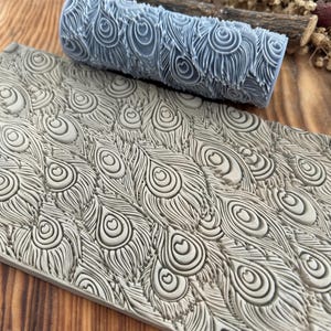 May include: A light gray textured roller and a matching rectangular tile, both featuring a peacock feather design. The roller is cylindrical, and the tile is rectangular. The design is a repeating pattern of stylized peacock feathers.