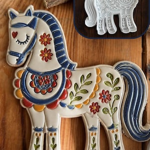 May include: A white clay horse figure with blue, red, and yellow painted floral details, featuring a closed eye and a heart on its cheek. An inset shows a white plastic clay cutter and stamp with the same horse shape and intricate embossed patterns.