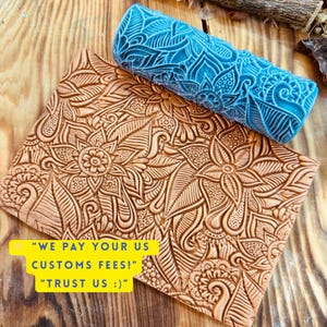 Op de afbeelding: Een bruin lederen ambachtelijk stuk met een bloemmotief en een blauwe roller met een bijpassend patroon. Het leren stuk heeft een vierkante vorm. De roller is cilindrisch. De afbeelding bevat de tekst "WE PAY YOUR US CUSTOMS FEES!" en "TRUST US :)".