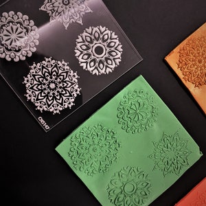 May include: A clear plastic sheet with four different mandala designs. The designs are white and are on a clear background. There are three silicone sheets with the same mandala designs in green, orange, and brown.