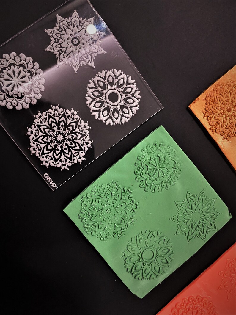 May include: A clear plastic sheet with four different mandala designs. The designs are white and are on a clear background. There are three silicone sheets with the same mandala designs in green, orange, and brown.