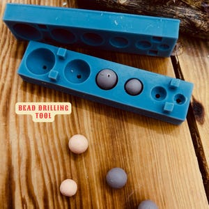 May include: Blue silicone mould with multiple round cavities for bead drilling. The mould is labelled "Bead Drilling Tool". There are four small beads, two beige and two grey, on a wooden surface.
