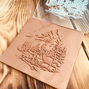 May include: A clear acrylic stamp featuring a design of a cabin in the woods with mountains in the background. The stamp is being used to create an impression on a brown leather surface.