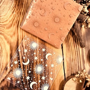 May include: Two clear acrylic stamps with a repeating pattern of suns, moons, and stars. The stamps are on a wooden surface.