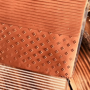 Könnte beinhalten: Nahaufnahme einer braunen Tonscheibe mit zwei verschiedenen Prägemustern. Das obere Muster ist eine Reihe horizontaler Linien. Das untere Muster ist ein sich wiederholendes geometrisches Muster aus kleinen Quadraten mit einem Punkt in der Mitte. Die Tonscheibe liegt auf einer durchsichtigen Plastikfolie mit einem Gittermuster.