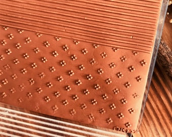 May include: A close-up of a brown clay sheet with two different embossed patterns. The top pattern is a series of horizontal lines. The bottom pattern is a repeating geometric design of small squares with a dot in the center. The clay sheet is on top of a clear plastic sheet with a grid pattern.
