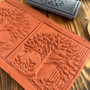May include: A close-up of a red clay tile with two raised impressions of trees. The tile is textured with lines and has a raised border. A blue silicone roller with a raised tree design is in the background.