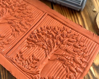 May include: A close-up of a red clay tile with two raised impressions of trees. The tile is textured with lines and has a raised border. A blue silicone roller with a raised tree design is in the background.