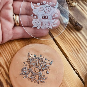 Puede incluir: Un sello de acrílico transparente con un diseño de una vaca de las Highlands con una corona de flores y corazones. El sello se está sosteniendo en una mano. El sello también se muestra sobre una superficie de cuero marrón.