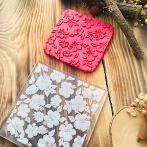 Könnte beinhalten: Zwei transparente Acrylstempel mit einem Blumenmuster. Der obere Stempel ist rot und der untere Stempel ist transparent mit weißen Blumen.