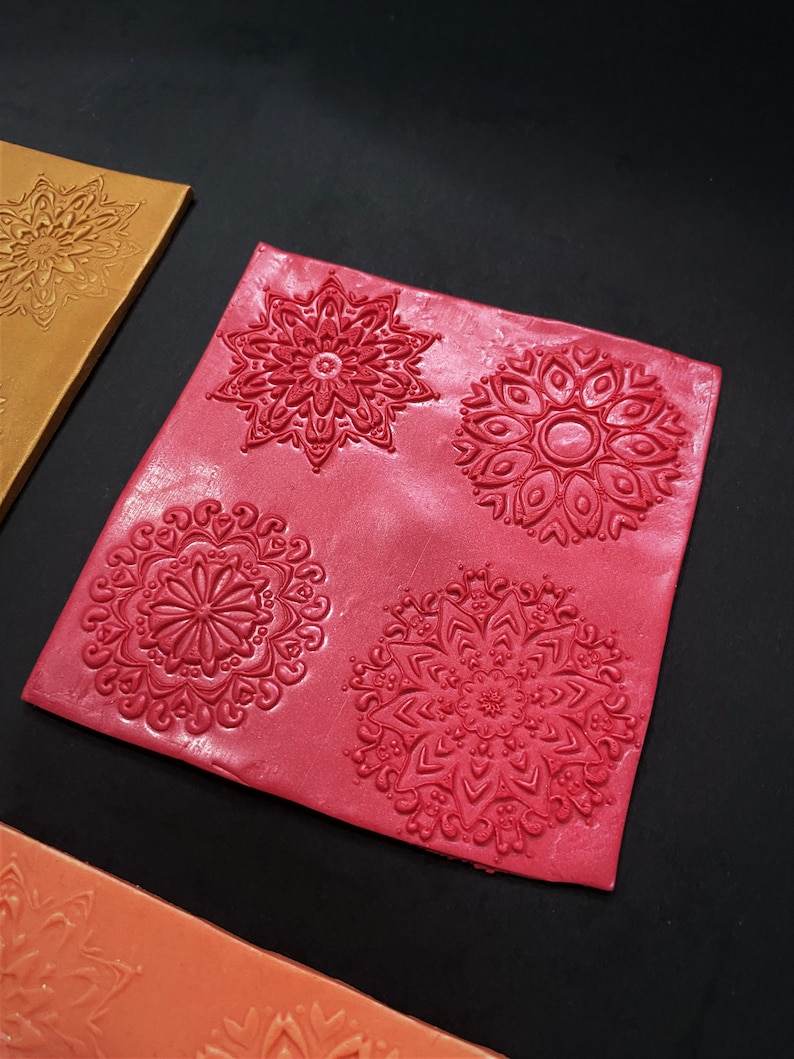 May include: Four red polymer clay stamps with intricate mandala designs. The stamps are arranged in a square pattern on a red background.