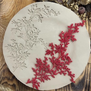 May include: A round, off-white ceramic mold with a floral design. One side features an embossed branch with leaves and berries, while the other side has a vibrant red, three-dimensional floral embellishment. The mold is set on a wooden surface.
