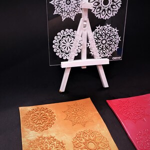 May include: Four clear acrylic stamps with intricate mandala designs. The stamps are displayed on a white easel. Two pieces of craft paper are shown with the mandala designs stamped in gold and red.
