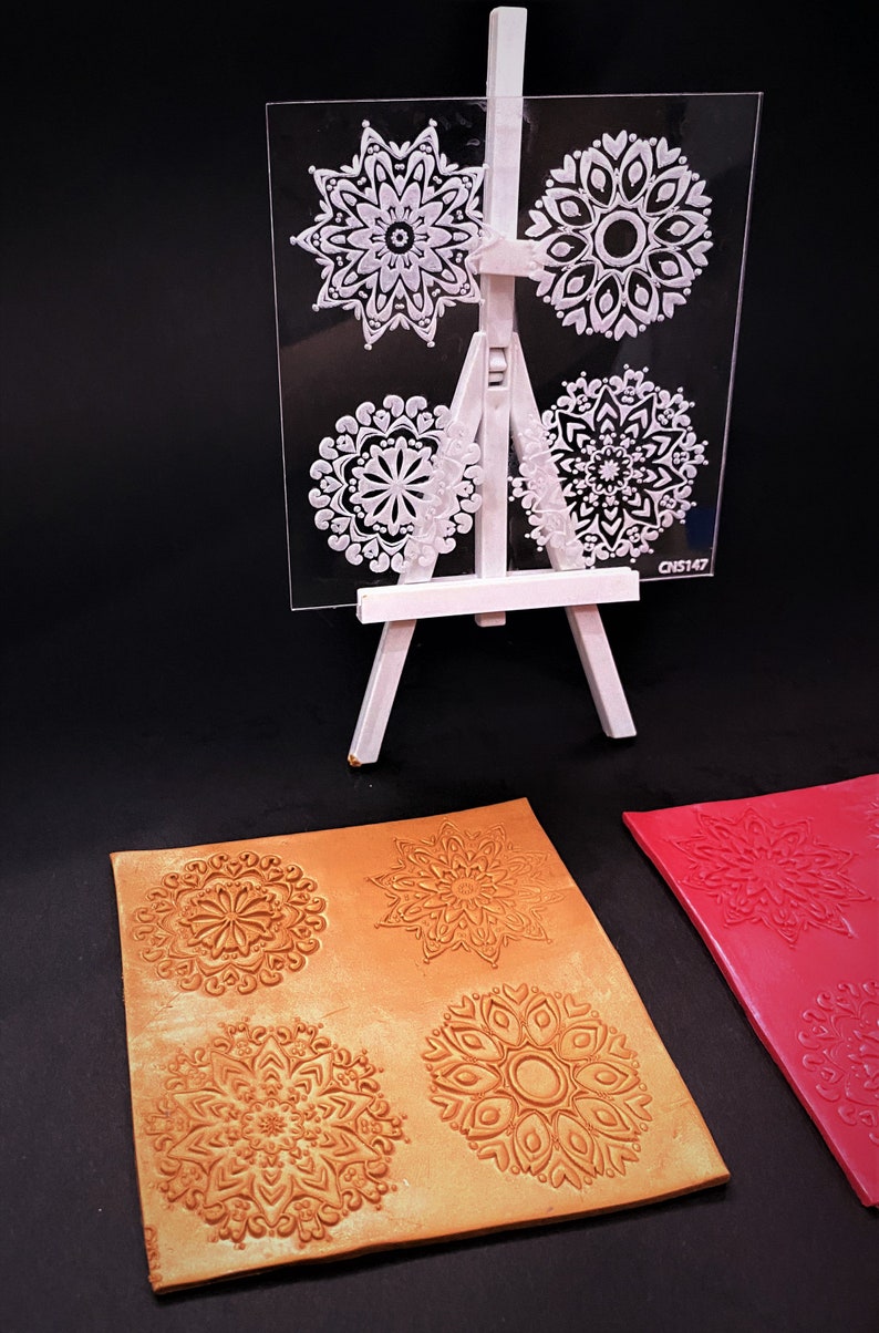 May include: Four clear acrylic stamps with intricate mandala designs. The stamps are displayed on a white easel. Two pieces of craft paper are shown with the mandala designs stamped in gold and red.