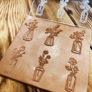 May include: A set of nine clear acrylic stamps featuring various floral designs in vases. The stamps are arranged on a brown clay surface.