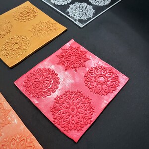 May include: Three pieces of clay with intricate mandala designs pressed into them. The clay is orange, red, and a darker orange. A clear plastic sheet with four mandala designs is also shown.