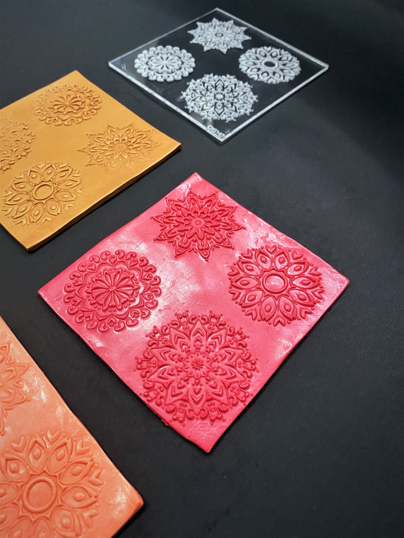 May include: Three pieces of clay with intricate mandala designs pressed into them. The clay is orange, red, and a darker orange. A clear plastic sheet with four mandala designs is also shown.