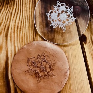 May include: A clear, round acrylic stamp with a floral design, next to a brown clay impression of the same floral pattern. The stamp and impression are on a wooden surface.