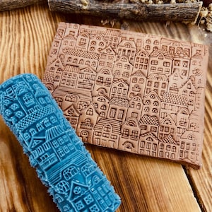 May include: A blue rolling pin and a brown rectangular block, both with detailed embossed cityscapes. The rolling pin is cylindrical, while the block is rectangular. The cityscape design includes houses, roofs, and windows.