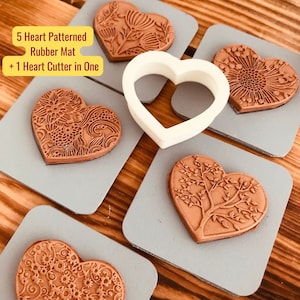 May include: Five heart-shaped rubber stamps with different intricate designs, including floral, tree, and geometric patterns. A white heart-shaped cookie cutter is also included. The stamps are on a gray surface.