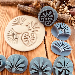 May include: A set of nine decorative stamps featuring various floral designs. The stamps are made of a light gray silicone material and are arranged on a wooden surface.
