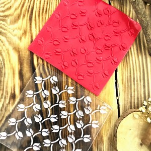 May include: A clear acrylic stamp with a repeating pattern of white tulips on a red background. The stamp is designed for crafting and scrapbooking.