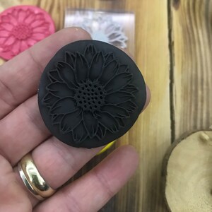 May include: A black rubber stamp with a detailed sunflower design. The stamp is being held in a hand with a gold ring.