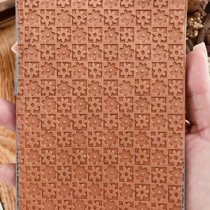 May include: A brown rubber stamp with a repeating pattern of small, raised flower designs. The stamp is rectangular and has a slightly textured surface.