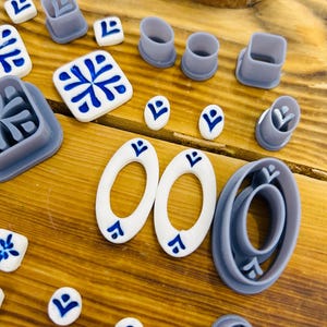 May include: A set of 14 blue and white ceramic tile-shaped cookie cutters, including a variety of geometric shapes and patterns. The cutters are arranged on a wooden surface.