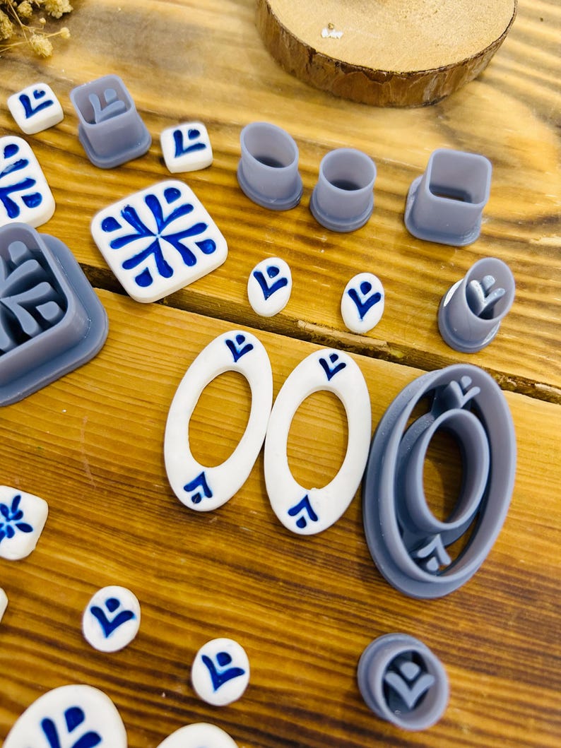 May include: A set of 14 blue and white ceramic tile-shaped cookie cutters, including a variety of geometric shapes and patterns. The cutters are arranged on a wooden surface.