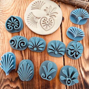 May include: A set of 10 blue polymer clay stamps with various floral and geometric designs. The stamps are arranged on a wooden surface. One of the stamps is a larger, beige polymer clay stamp with three different designs.