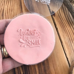 May include: A pink silicone stamp with the text "Wake up and smile" in a white, stylized font. The stamp is in a clear plastic case.