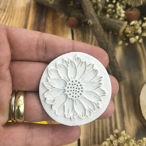 Peut inclure: Moule en silicone blanc avec un motif en relief de tournesol. Le moule est rond et a une surface lisse.