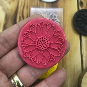 May include: A red round silicone stamp with a detailed sunflower design. The sunflower has a textured center and petals.