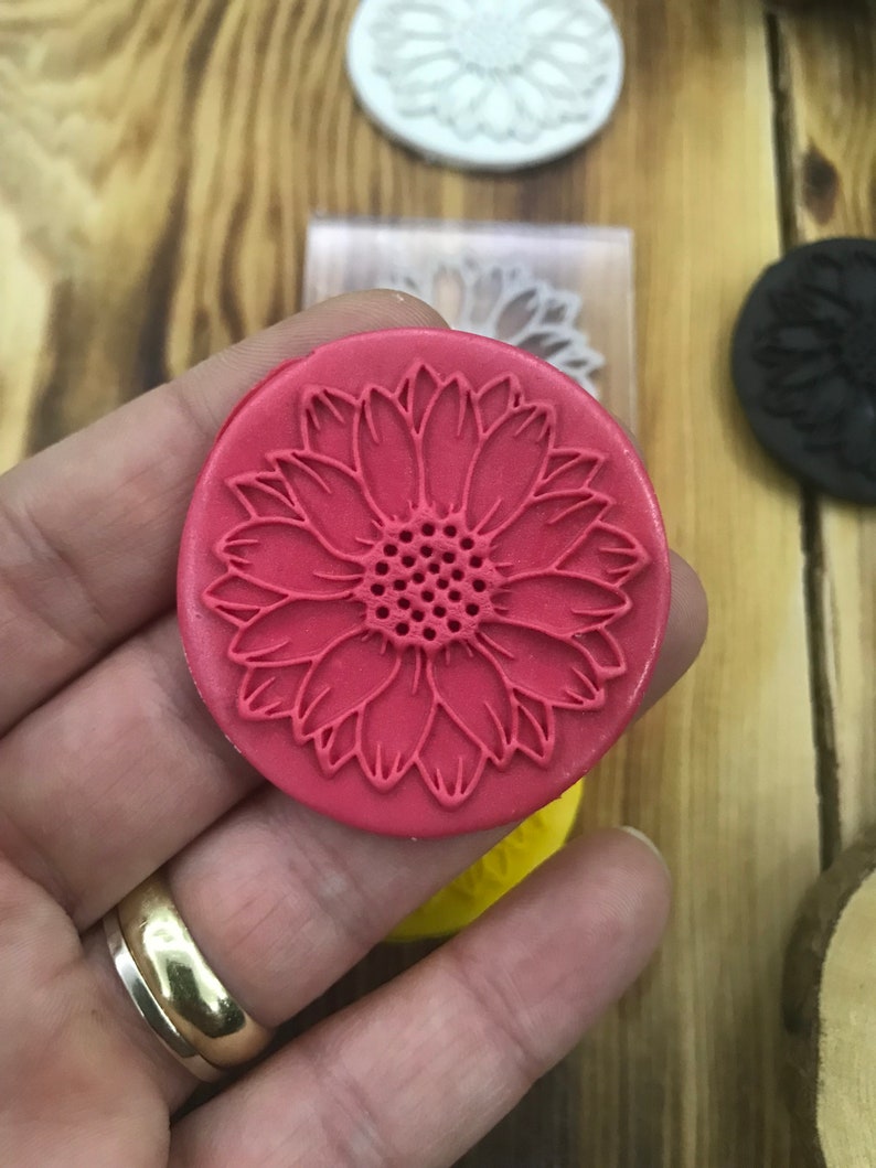May include: A red round silicone stamp with a detailed sunflower design. The sunflower has a textured center and petals.