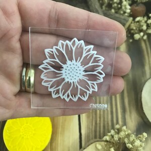 May include: Clear acrylic stamp with a white sunflower design. The stamp is approximately 2 inches by 2 inches and has the code CNS099 printed on the bottom.