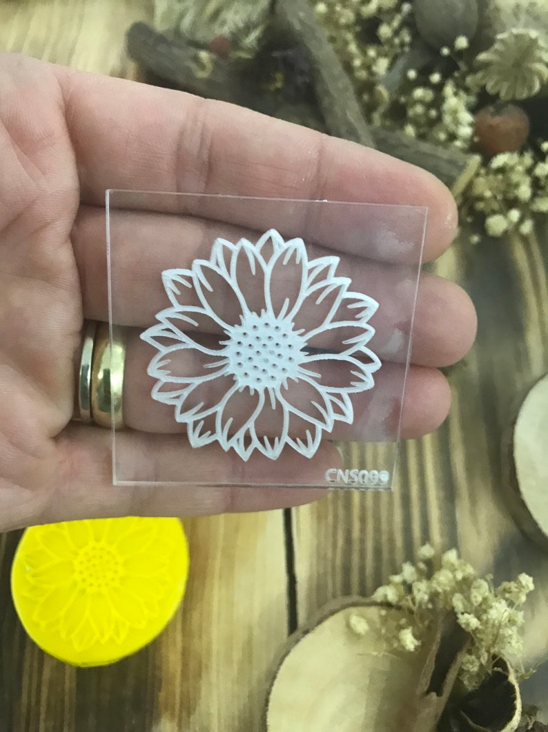 May include: Clear acrylic stamp with a white sunflower design. The stamp is approximately 2 inches by 2 inches and has the code CNS099 printed on the bottom.