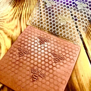 Puede incluir: Una lámina de plástico transparente con un patrón de panal y abejas impresas en ella. La lámina se coloca sobre una baldosa de arcilla marrón con un patrón de panal similar y abejas en relieve en la superficie.