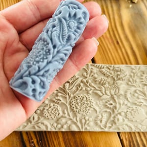 Puede incluir: Un sello cilíndrico azul claro con un patrón floral se sostiene en una mano. El sello tiene tallas en relieve detalladas de flores y hojas. Un sello beige a juego con un diseño floral similar está en el fondo.