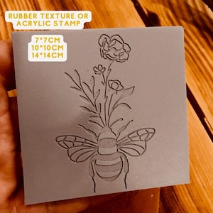 Op de afbeelding: Een grijze rubberen of acryl stempel met een bij- en bloemenontwerp. De stempel is verkrijgbaar in de maten 7x7cm, 10x10cm of 14x14cm.
