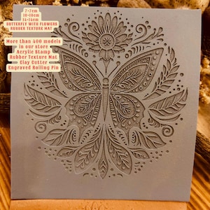May include: A grey rubber texture mat featuring an intricate design of a butterfly with flowers. The mat measures 7-7cm, 10-10cm, and 14-14cm. The text "More than 400 models in our store Acrylic Stamp Rubber Texture Mat Clay Cutter Engraved Rolling Pin" is printed on the mat.