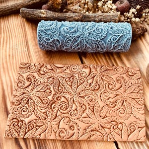 Puede incluir: Un rodillo azul estampado y una pieza rectangular marrón con un diseño de pulpo. El rodillo es cilíndrico y presenta un patrón repetitivo de tentáculos de pulpo. La pieza rectangular tiene un diseño en relieve similar. Los artículos están sobre una superficie de madera.