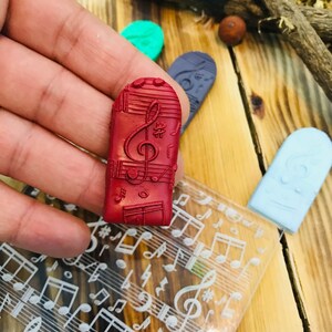 K&ouml;nnte beinhalten: Ein roter rechteckiger Stempel mit einem Noten-Design, gehalten in einer Hand. Weitere Stempel in Gr&uuml;n, Blau und Hellblau sind sichtbar. Eine transparente Folie mit Noten ist ebenfalls vorhanden. Der Hintergrund ist eine Holzoberfl&auml;che.