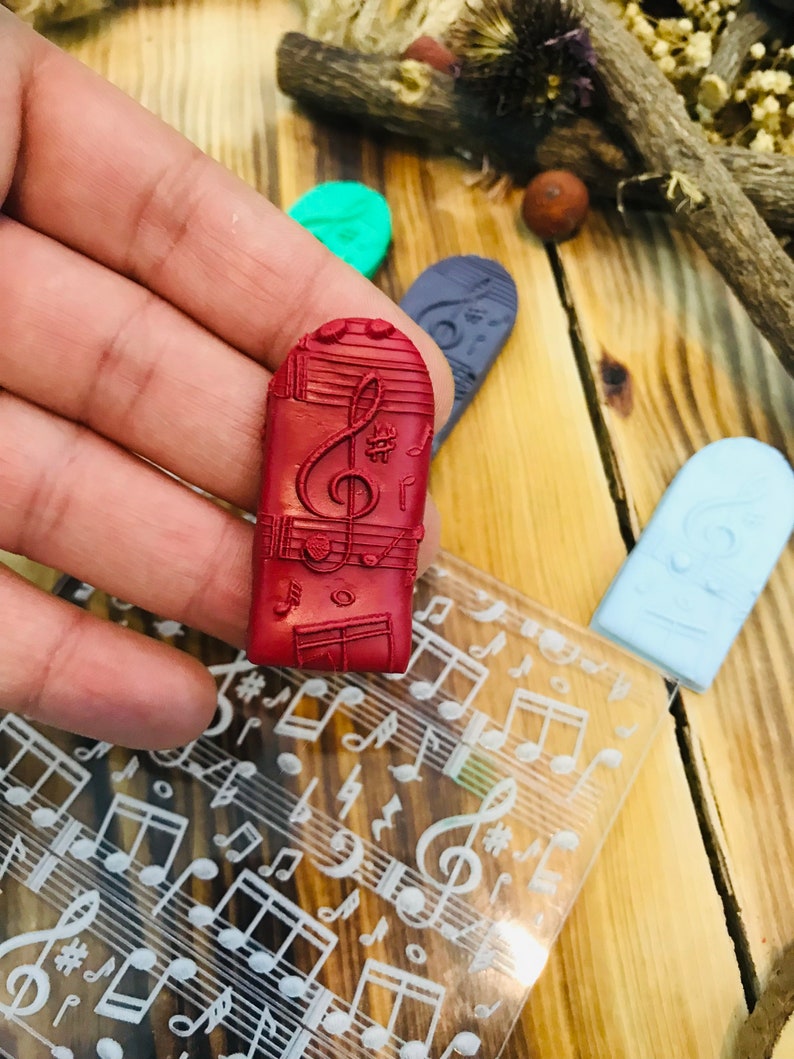 K&ouml;nnte beinhalten: Ein roter rechteckiger Stempel mit einem Noten-Design, gehalten in einer Hand. Weitere Stempel in Gr&uuml;n, Blau und Hellblau sind sichtbar. Eine transparente Folie mit Noten ist ebenfalls vorhanden. Der Hintergrund ist eine Holzoberfl&auml;che.